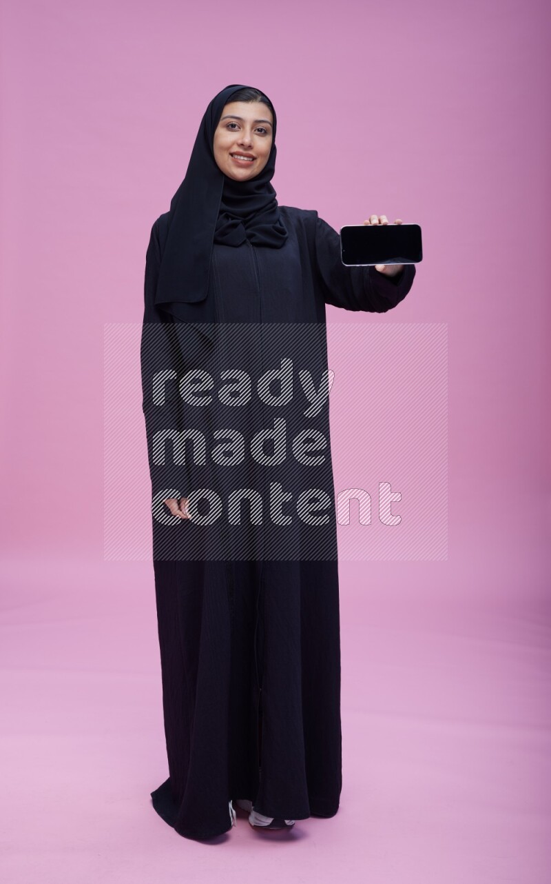 Saudi woman wearing Abaya standing showing phone to camera on pink background