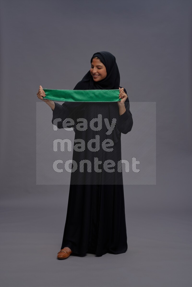 Saudi woman wearing Abaya standing holding Saudi flag on gray background