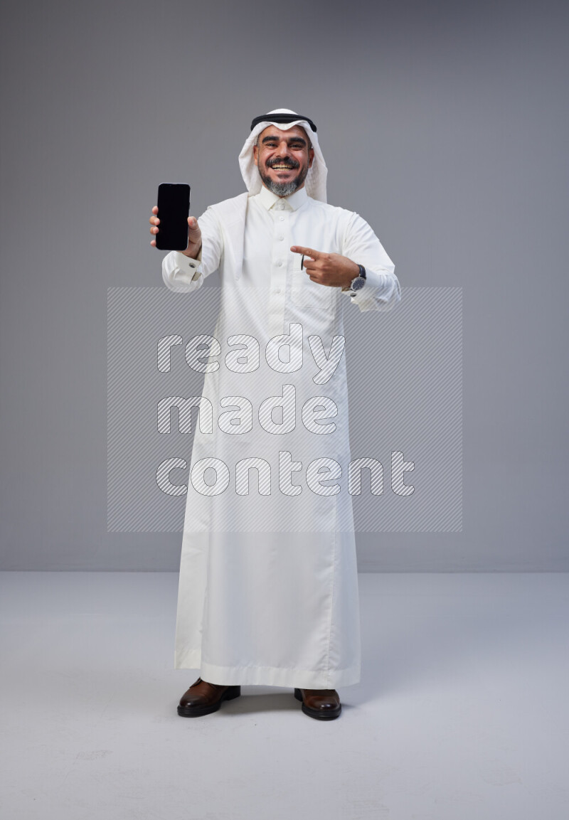 Saudi man Wearing Thob and white Shomag standing showing phone to camera on Gray background