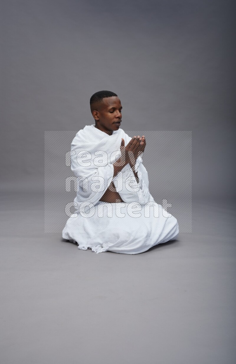A man wearing Ehram sitting on floor dua'a on gray background