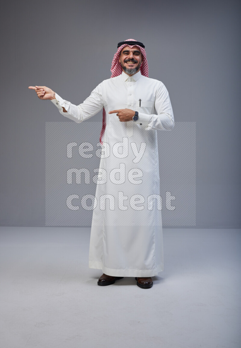 Saudi man Wearing Thob and red white Shomag standing interacting with the camera on Gray background