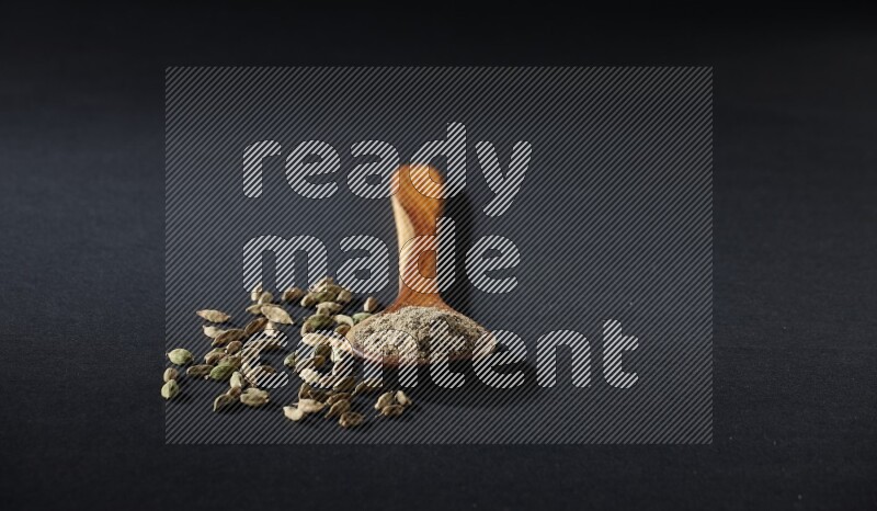 A wooden ladle full of cardamom powder and cardamom seeds beside it on black flooring