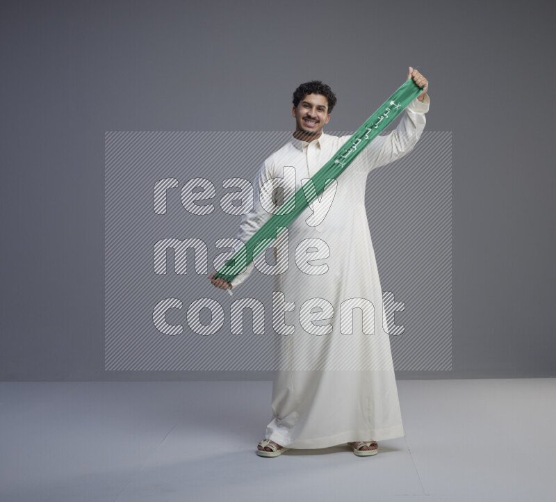 A Saudi man standing wearing thob holding Saudi flag scarf on gray background