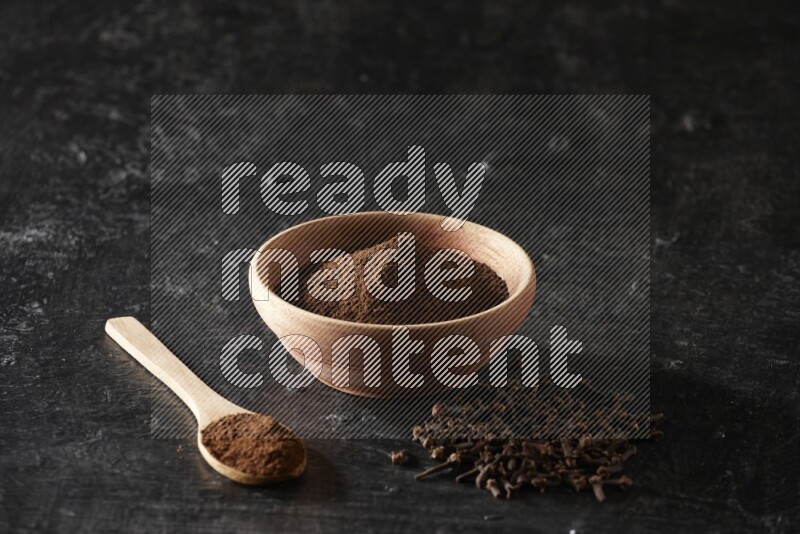 A wooden bowl and a wooden spoon full of cloves powder with spreaded cloves on a textured black flooring