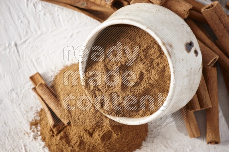 Ceramic beige bowl over filled with cinnamon powder and cinnamon sticks around the bowl on a textured white background in different angles
