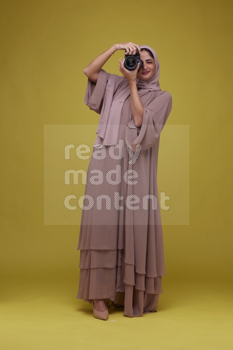 A woman Holding a Camera on a Yellow Background wearing Brown Abaya with Hijab
