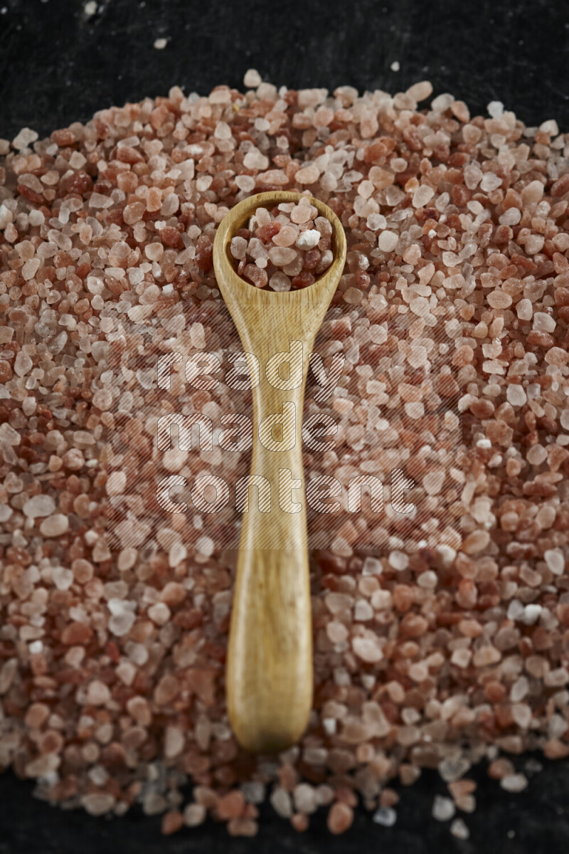 A wooden spoon full of coarse himalayan salt crystals on a bunch of the crystals on black background