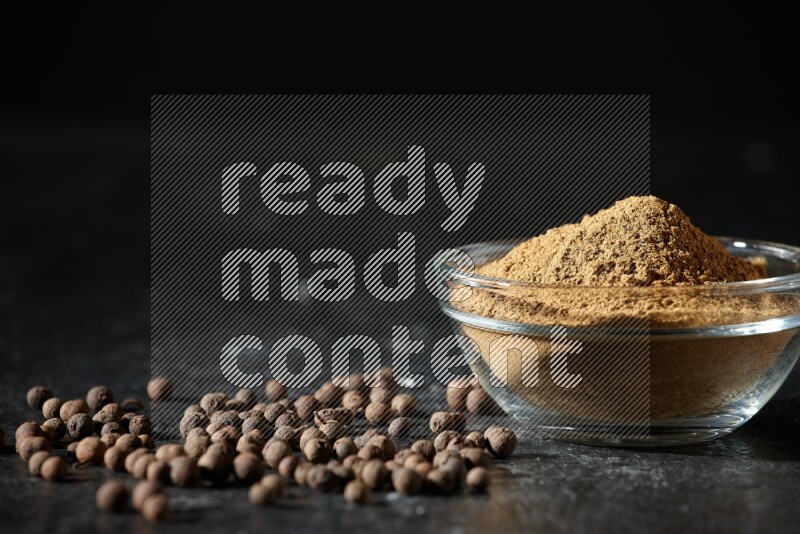 A glass bowl full of allspice powder and whole balls spreaded on a black flooring