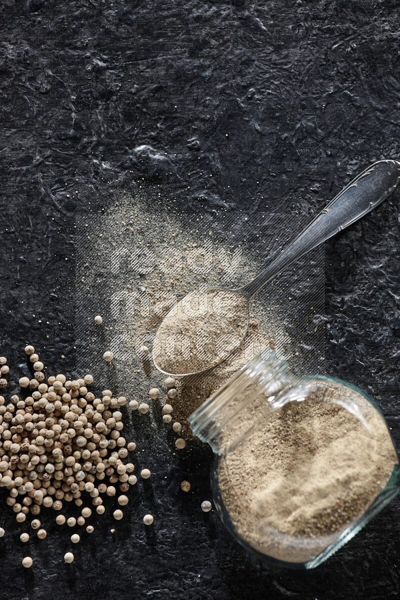 A flipped herbal glass jar and metal spoon full of white pepper powder with spilled powder and pepper beads on textured black flooring