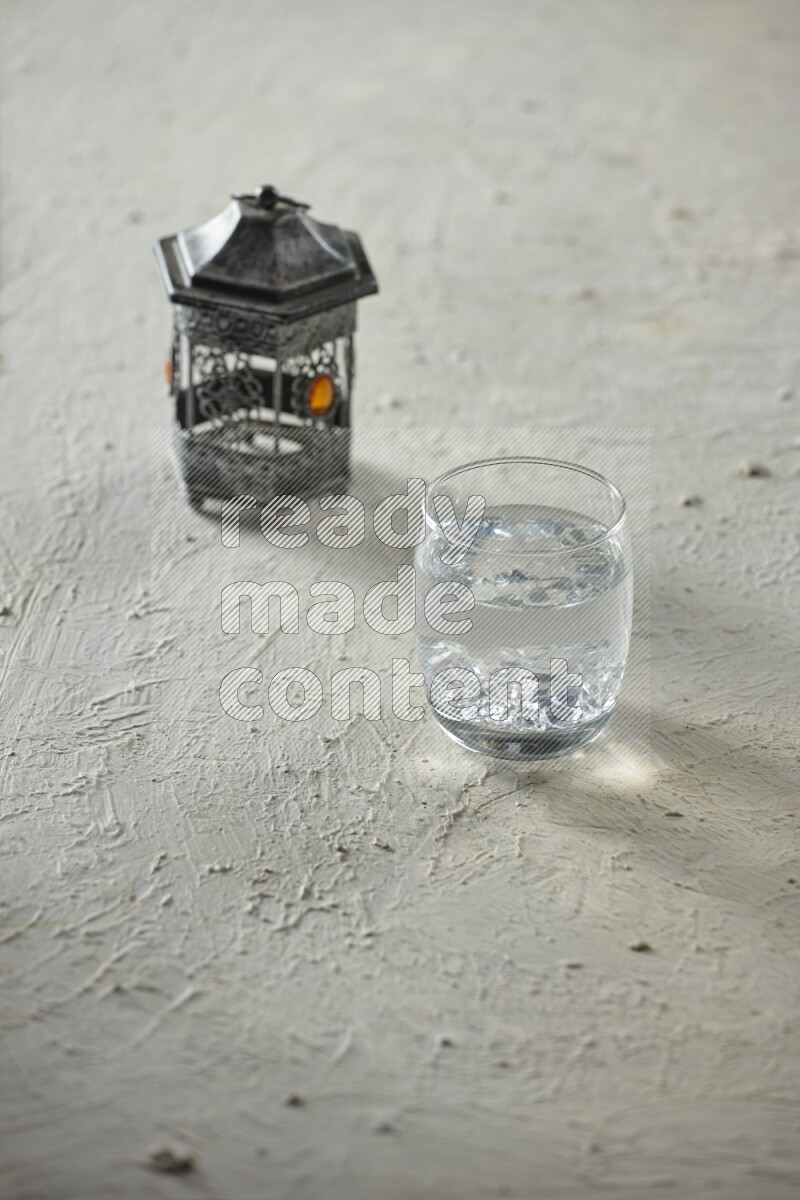 A silver lantern with different drinks, dates, nuts, prayer beads and quran on textured white background