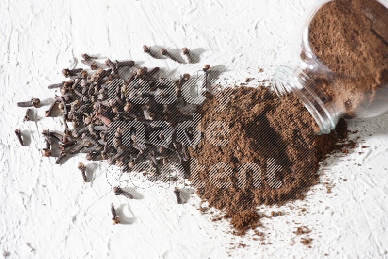 وعاء زجاجي للتوابل مقلوب ممتلئ ببودرة القرنفل وتناثرت البودرة منه مع حبوب القرنفل المنتشرة على أرضية بيضاء