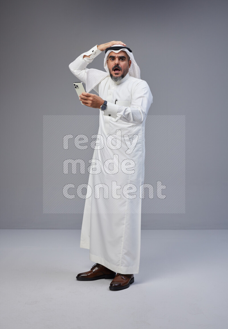 Saudi man Wearing Thob and white Shomag standing texting on phone on Gray background