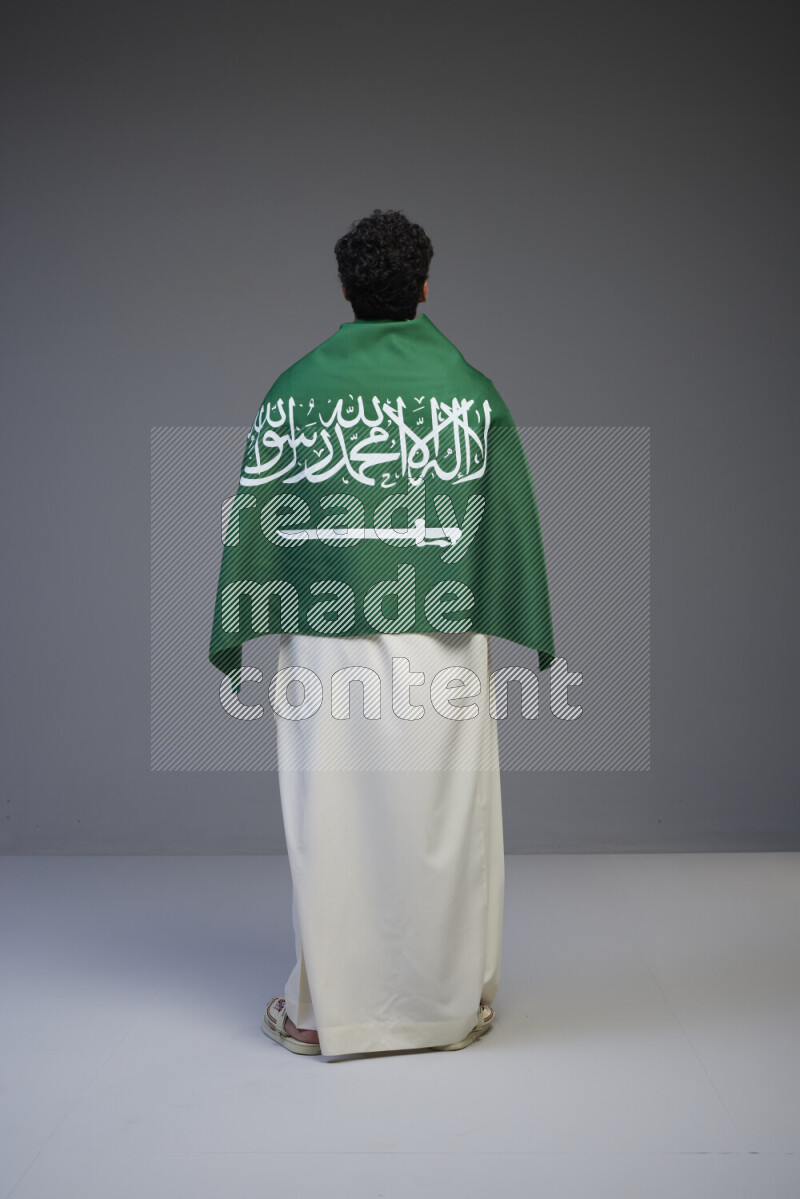 A Saudi man standing wearing thob wrapping big Saudi flag on gray background