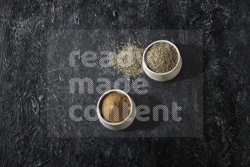 2 beige bowls, one full of cumin powder and the other full of seeds on a textured black flooring