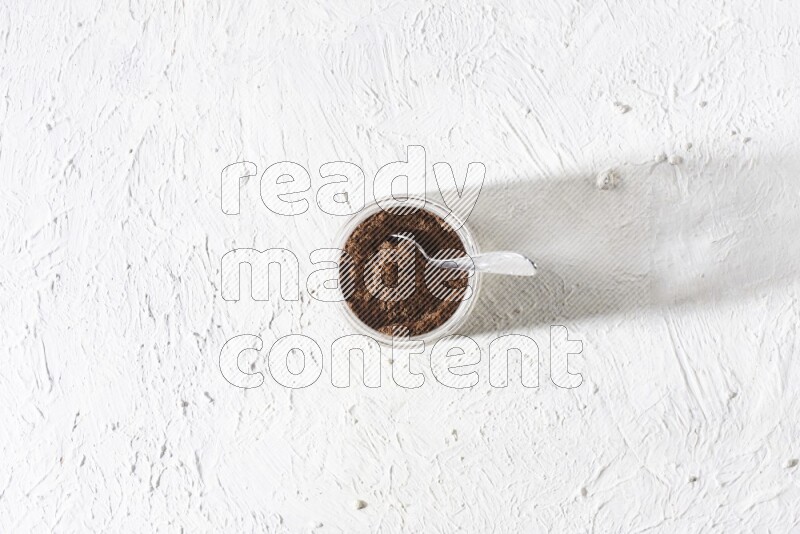 A glass jar full of cloves powder with a metal spoon on a textured white flooring