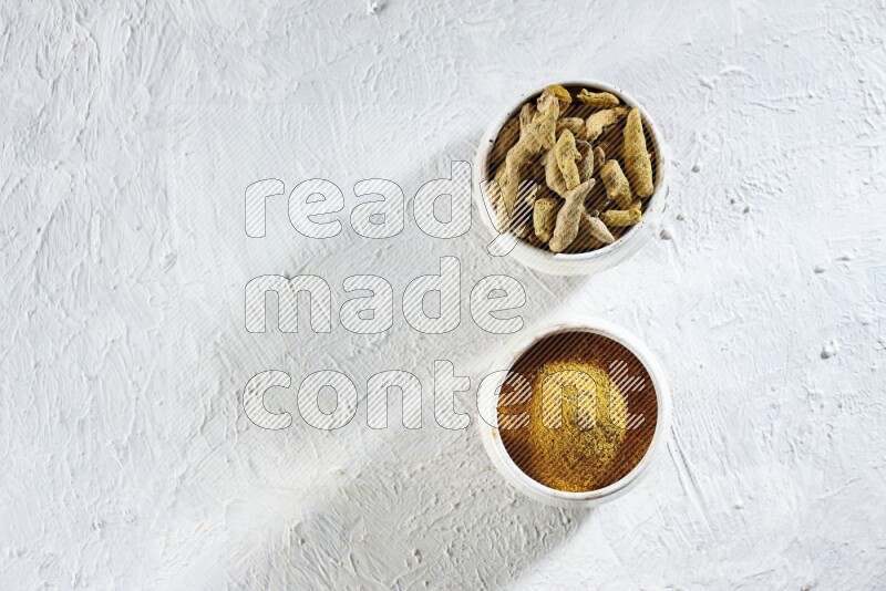 وعاءان فخاريان بيج ممتلئان ببودرة الكركم وأصابع الكركم المجففة على خلفية بيضاء