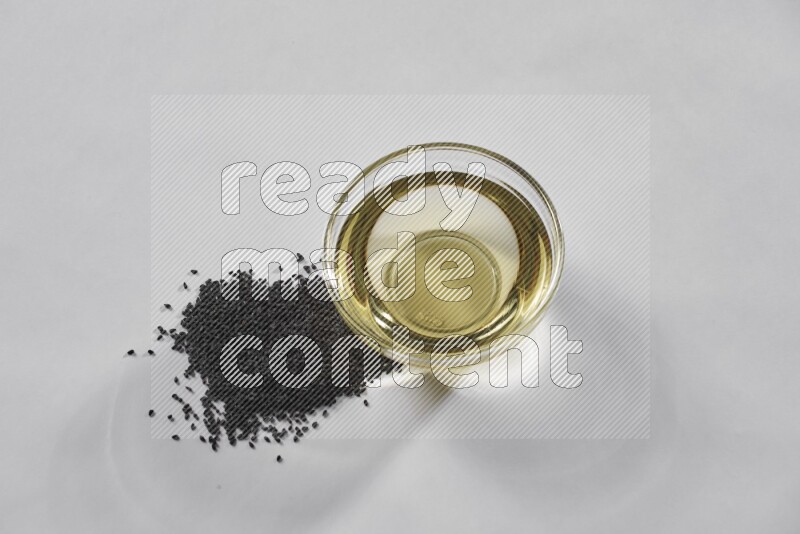 A glass bowl full of black seeds oil with black seeds beside it on a white flooring