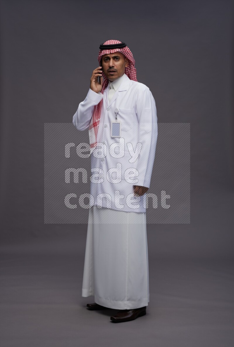 Saudi man wearing thob with lab coat and shomag with pocket employee badge standing talking on phone on gray background