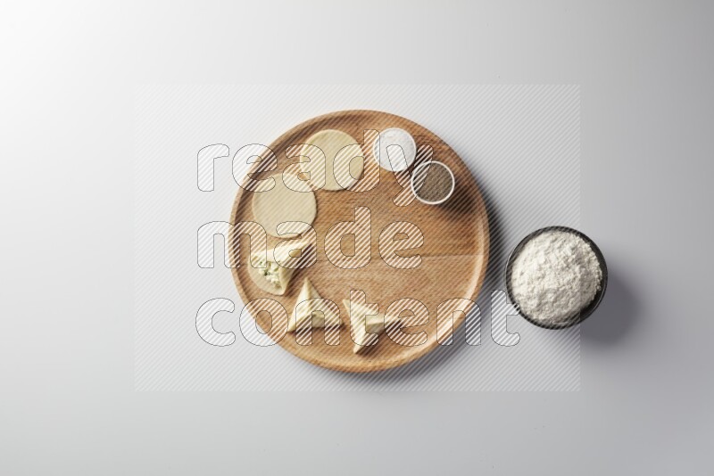 two closed sambosas and one open sambosa filled with cheese while flour, salt, and black pepper aside in a wooden dish on a white background
