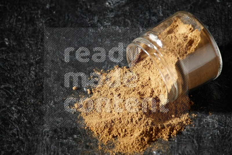 A flipped glass jar full of allspice powder and powder spilled out of it on a textured black flooring