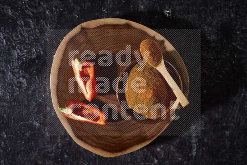 A wooden bowl full of ground paprika powder and sliced red bell pepper beside it, all on a wooden tray on black background