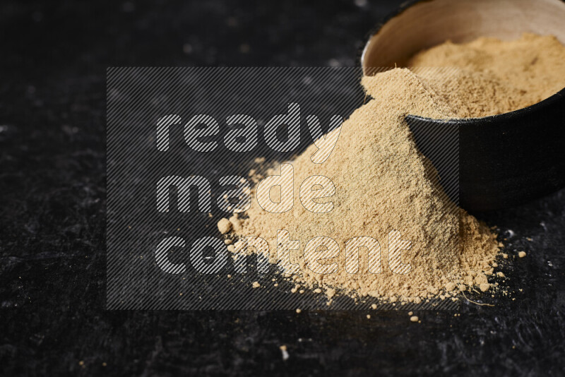 A black pottery bowl full of ground ginger powder with fallen powder from it on black background