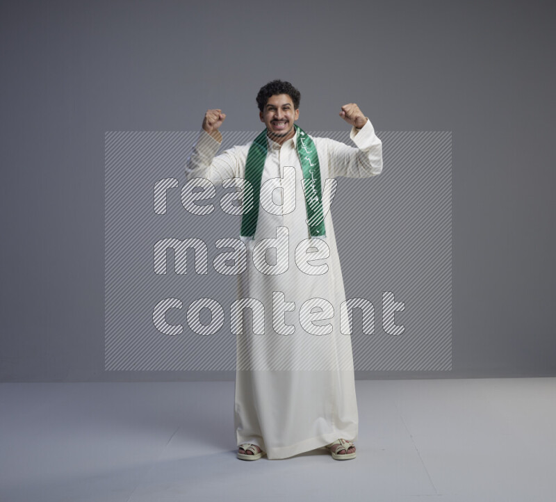 A Saudi man standing wearing thob and Saudi flag scarf on gray background