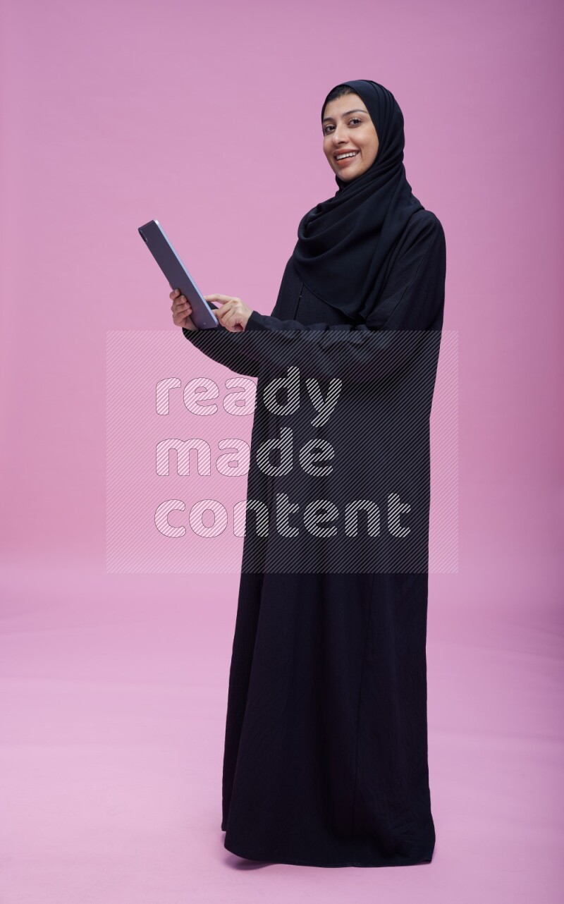 Saudi woman wearing Abaya standing working on tablet on pink background