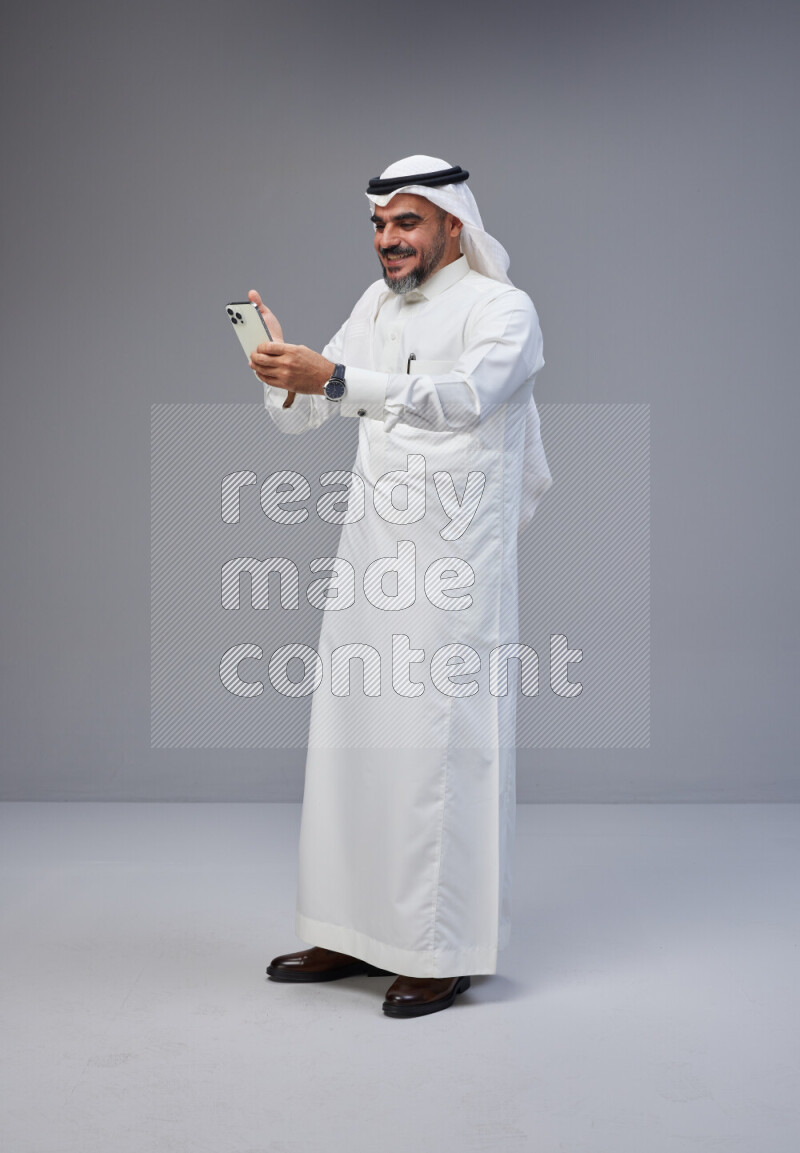 Saudi man Wearing Thob and white Shomag standing texting on phone on Gray background