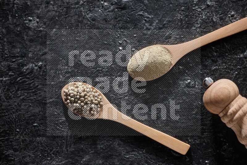 2 wooden spoons one full of white pepper powder and the other with pepper beads and a wooden pepper mill on textured black flooring