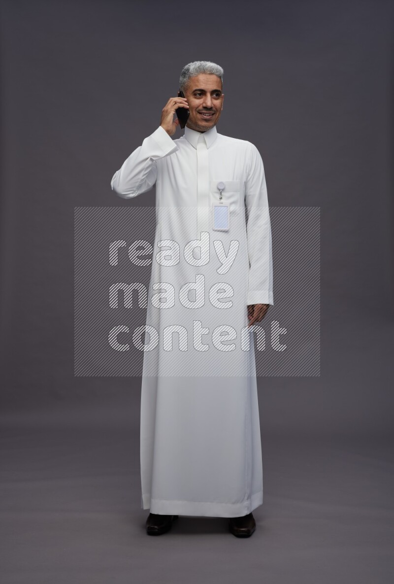 Saudi man wearing thob with pocket employee badge standing talking on phone on gray background