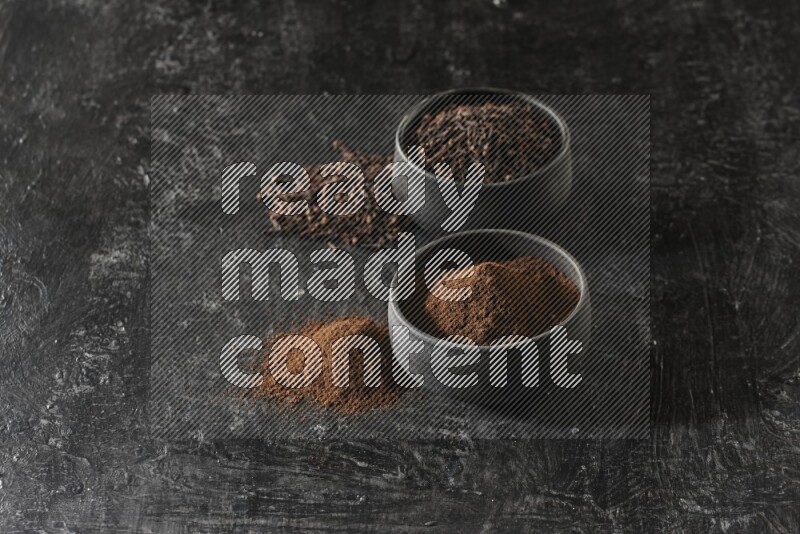 2 Black pottery bowls full of cloves and the other full of cloves powder on textured black flooring
