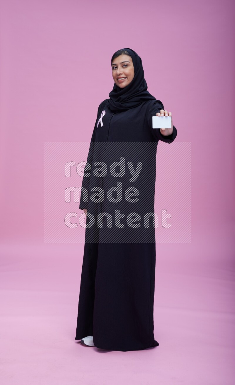 Saudi woman wearing pink ribbon on Abaya standing holding ATM card on pink background