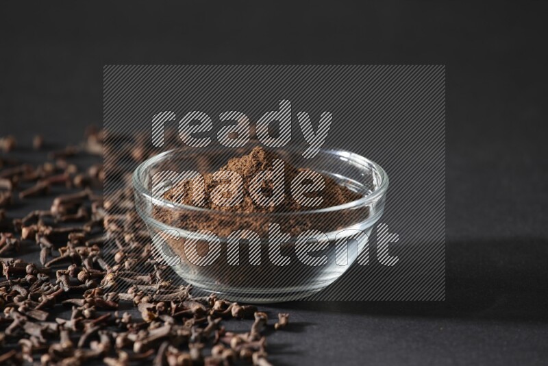 A glass bowl full of cloves powder with cloves grains spread on black flooring