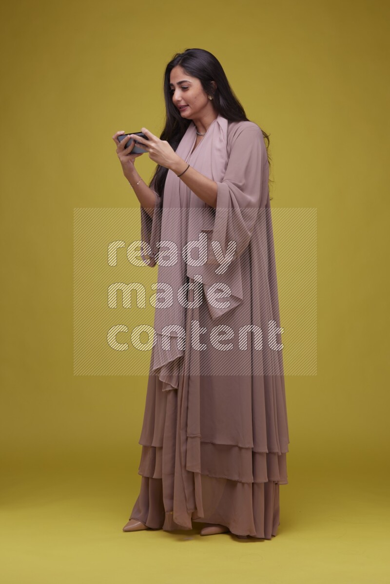 A woman Playing a Game on her smartphone on a Yellow Background wearing Brown Abaya