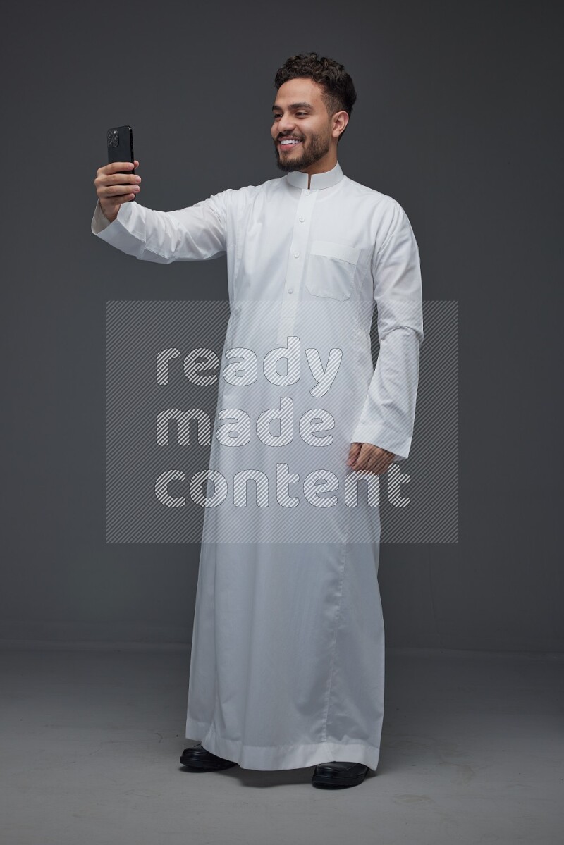 A Saudi man wearing Thobe and taking selfie with his phone making different poses eye level on a gray background