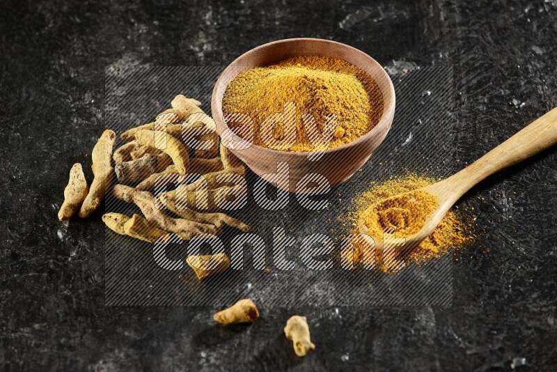 A wooden bowl and a wooden spoon full of turmeric powder with dried turmeric fingers on textured black flooring