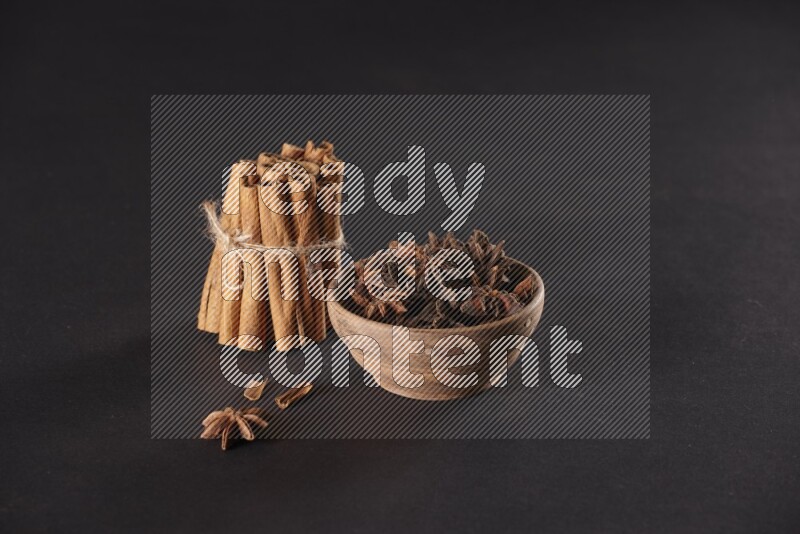 A stacked and bounded cinnamon sticks and a wooden bowl full of star anise on a black background