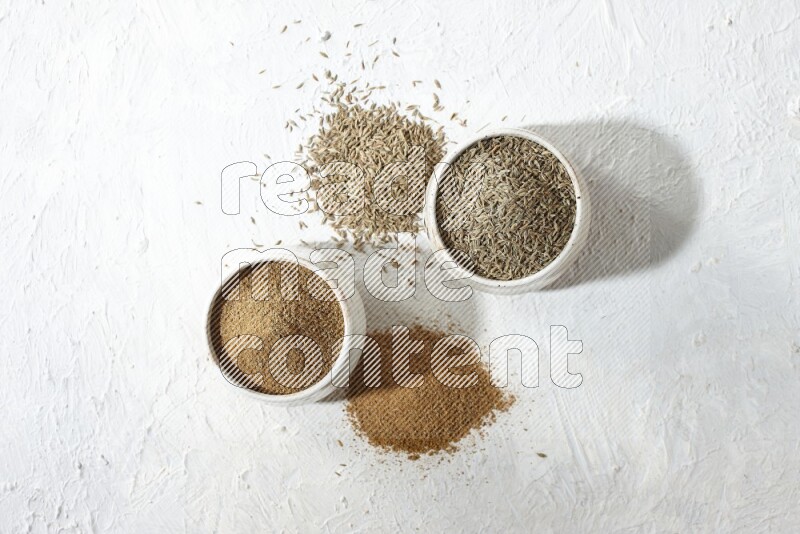 2 beige bowls full of cumin seeds and powder with spilled powder and seeds on textured white flooring