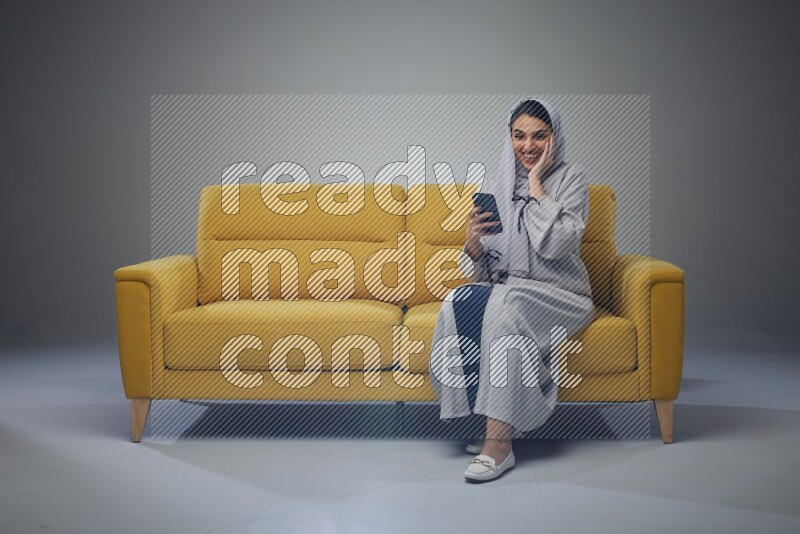 A Saudi woman wearing a light gray Abaya and head scarf setting on a yellow sofa and holding her phone while showing it's screen eye level on a grey background