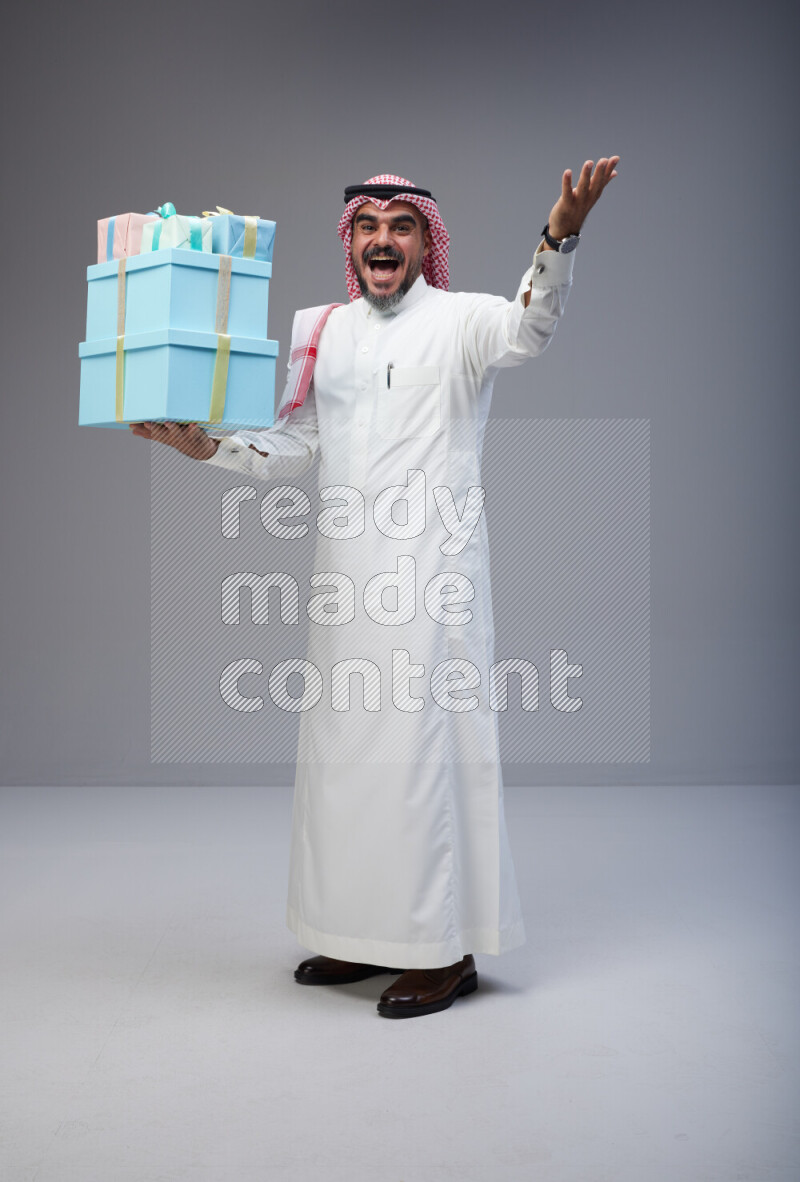 Saudi man Wearing Thob and red Shomag standing holding gift box on Gray background