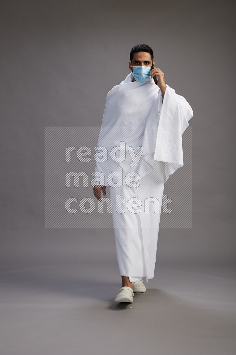 A man wearing Ehram with face mask Standing talking on phone on gray background