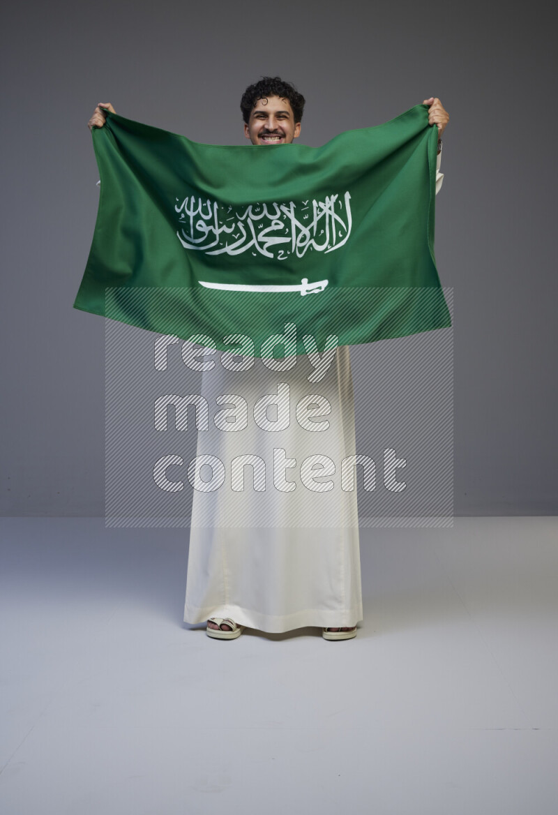 A Saudi man standing wearing thob holding big Saudi flag on gray background
