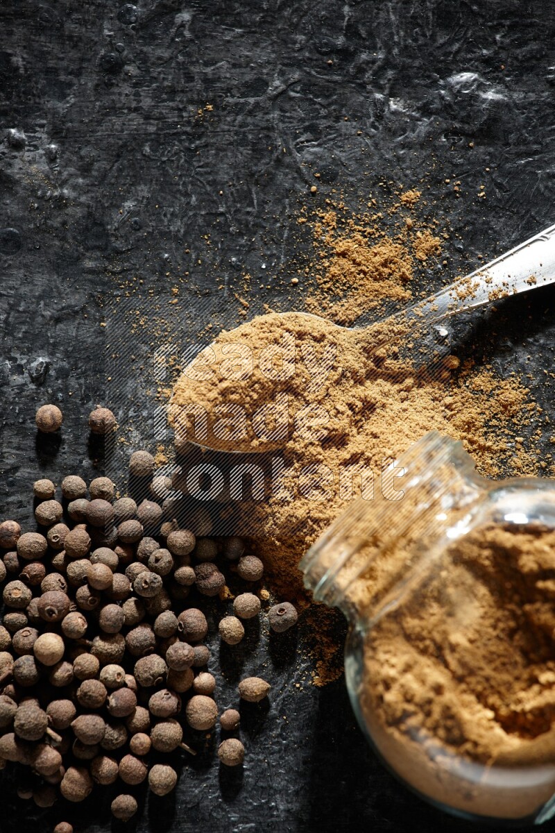 وعاء زجاجي للتوابل مقلوب وملعقة معدنية ممتلئان ببودرة بهار الفلفل الحلو والبودرة تناثرت من الوعاء مع بعض الحبوب على خلفية سوداء