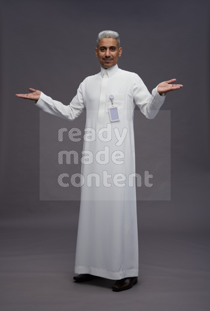 Saudi man wearing thob with pocket employee badge standing interacting with the camera on gray background