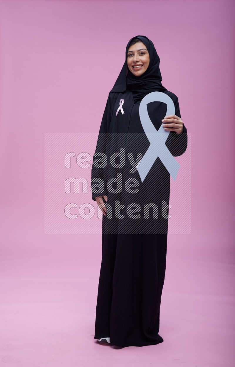 Saudi woman wearing pink ribbon on abaya standing holding awareness ribbon on plain pink background