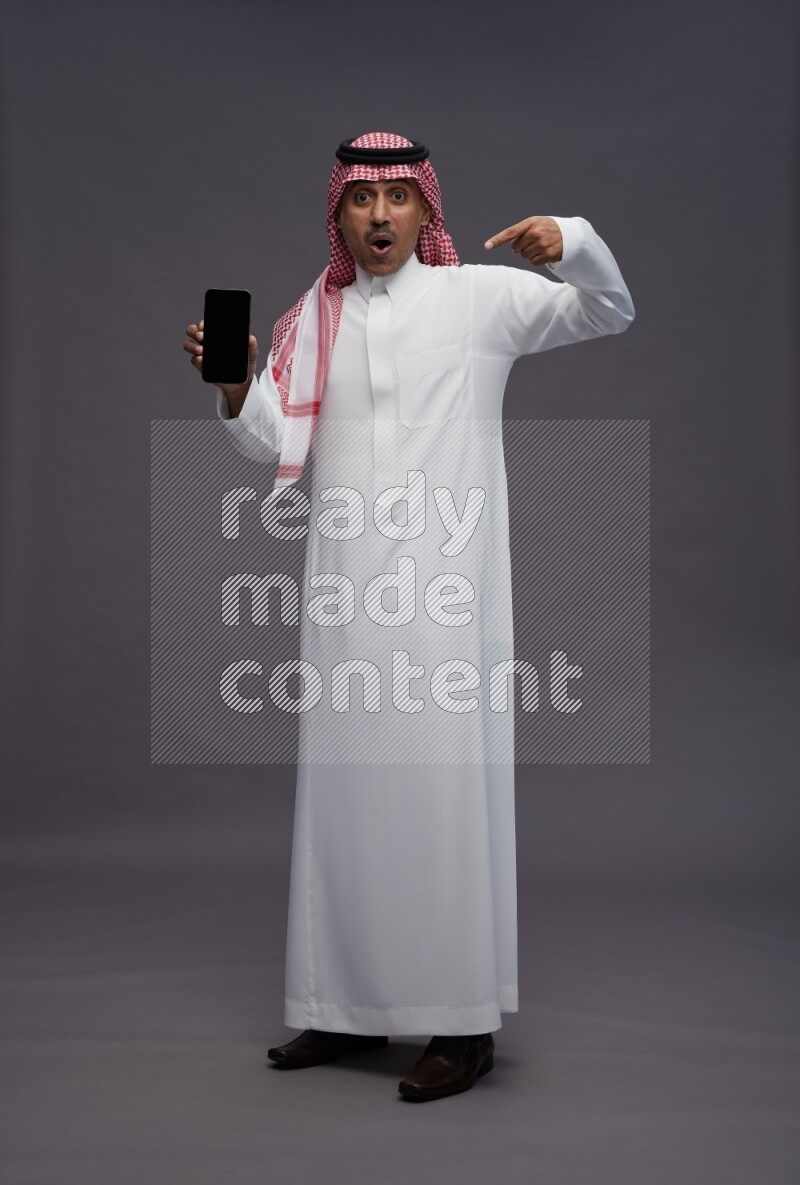 Saudi man wearing thob and shomag standing showing phone to camera on gray background