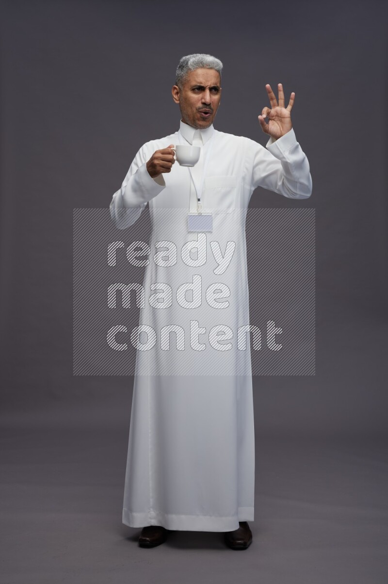 Saudi man wearing thob with neck strap employee badge standing holding mug on gray background