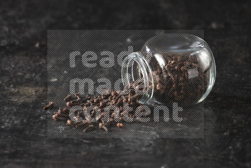 A flipped glass spice jar full of cloves and cloves came out of it on textured black flooring