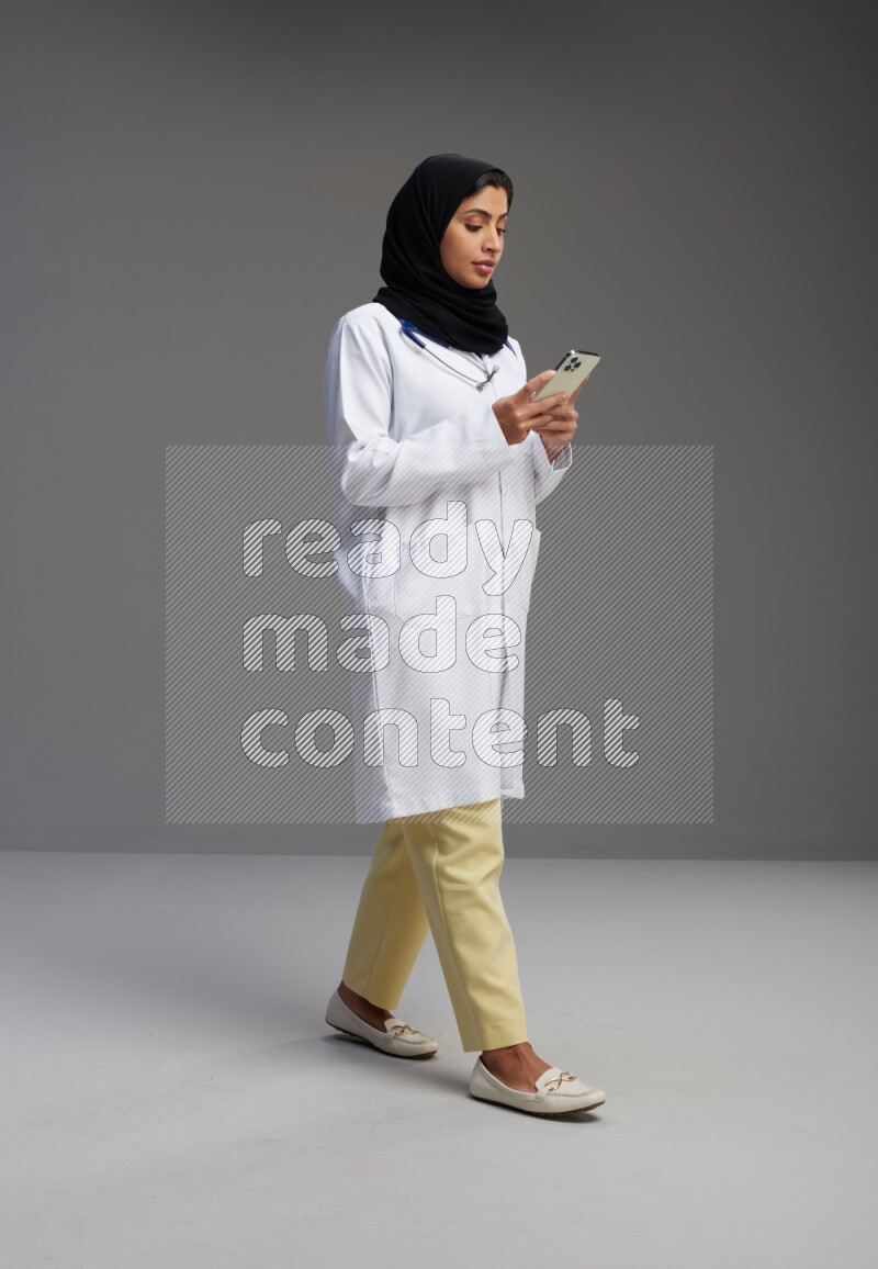 Saudi woman wearing lab coat with stethoscope standing texting on phone on Gray background